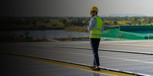 A man in a safety vest and hard hat holding a tablet walks along a field of solar panels.